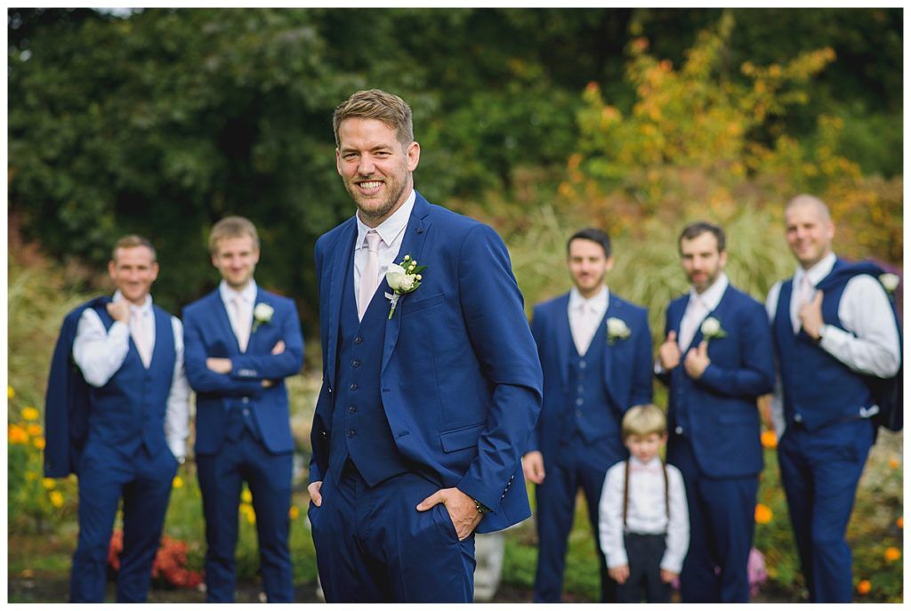 Groom smiles, posing with groomsmen in blue suits outdoors.