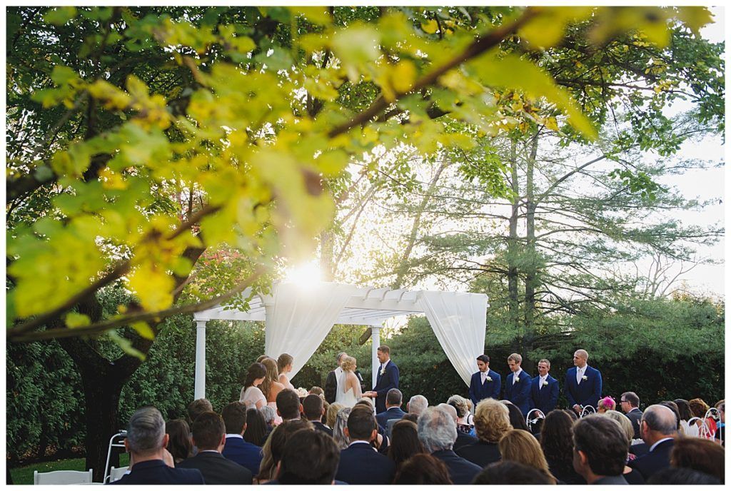 Wedding ceremony under a white arbor. Couple stands at altar, guests seated, sunlight streaming through trees.