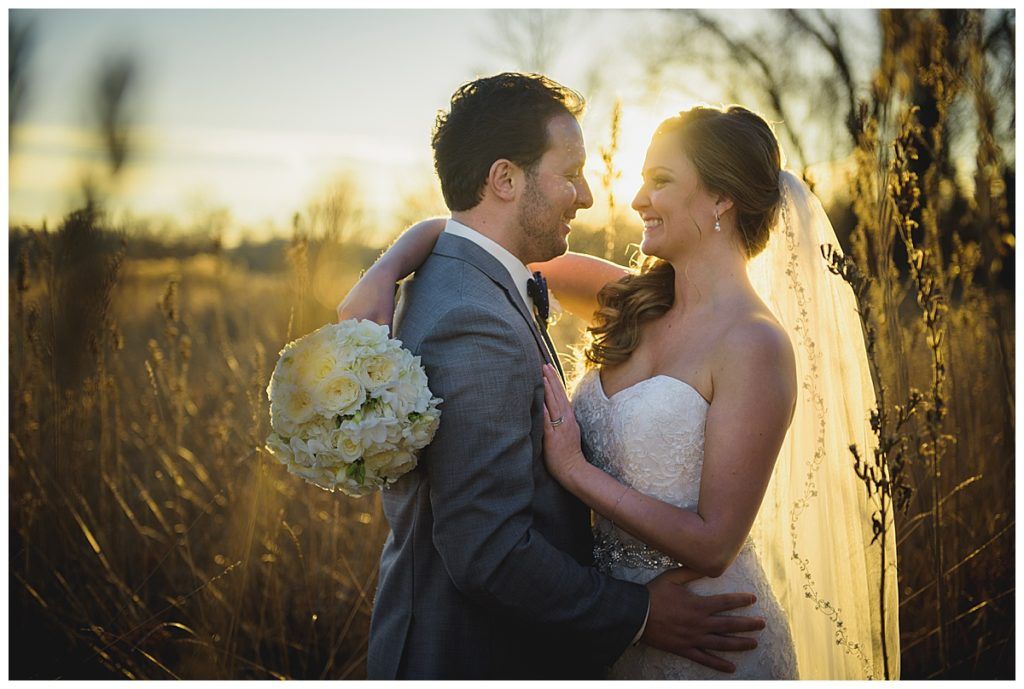 Newlyweds embrace in a field at sunset; bride in white gown, holding bouquet, groom in gray suit.