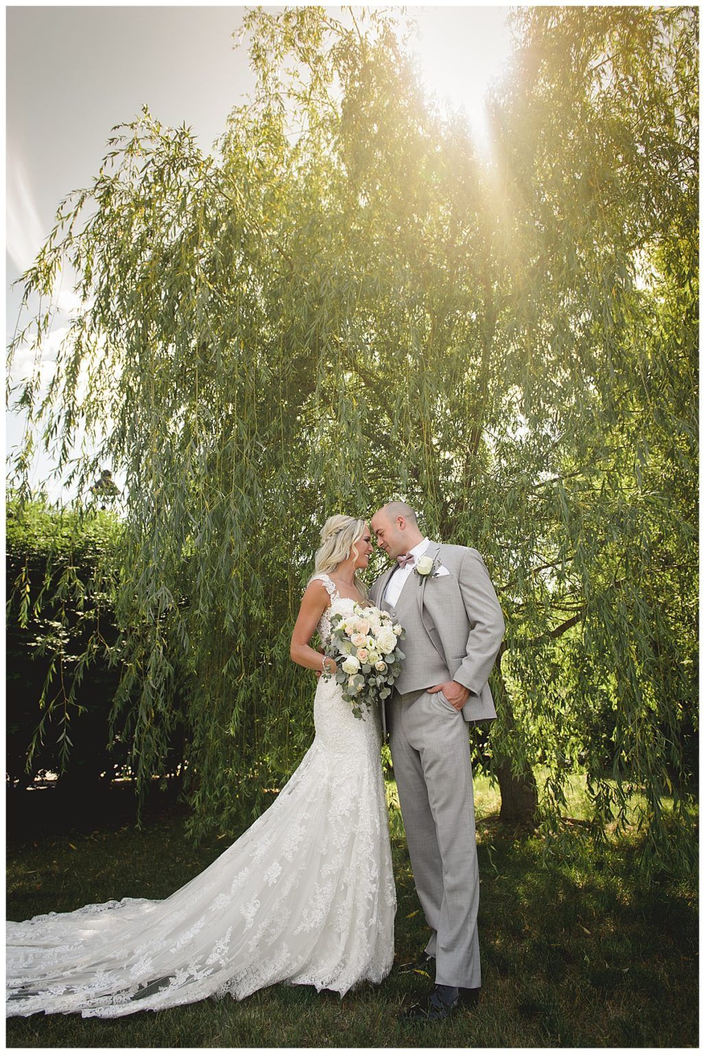 Bride and groom in wedding attire pose under a tree, sunlight shining through.