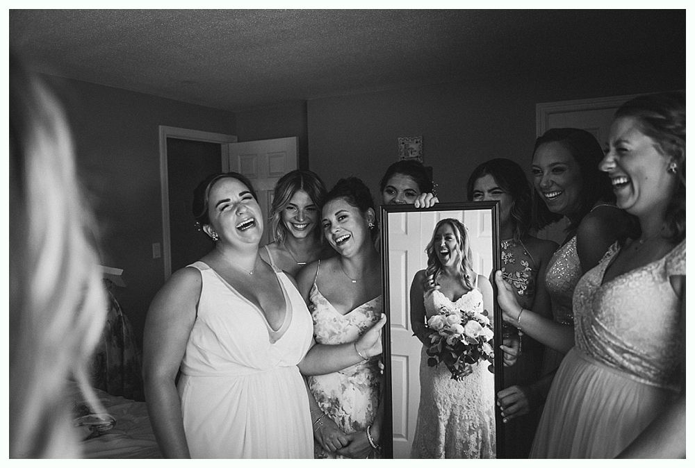Bridesmaids laugh with bride, who holds a mirror. They are in a room, celebrating a wedding. Black and white.
