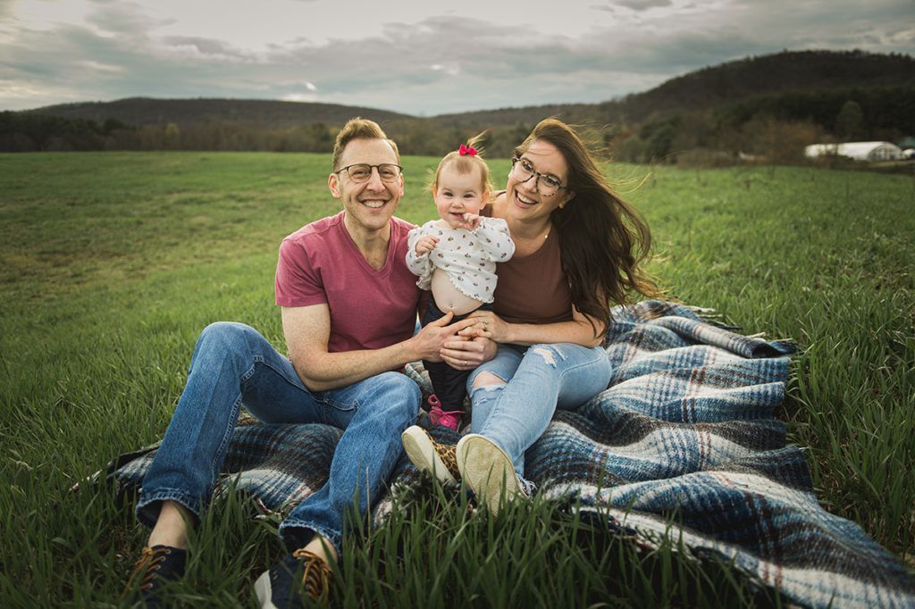 Family of three smiling while sitting on a blanket in a field. Cloudy sky with hills in the background.