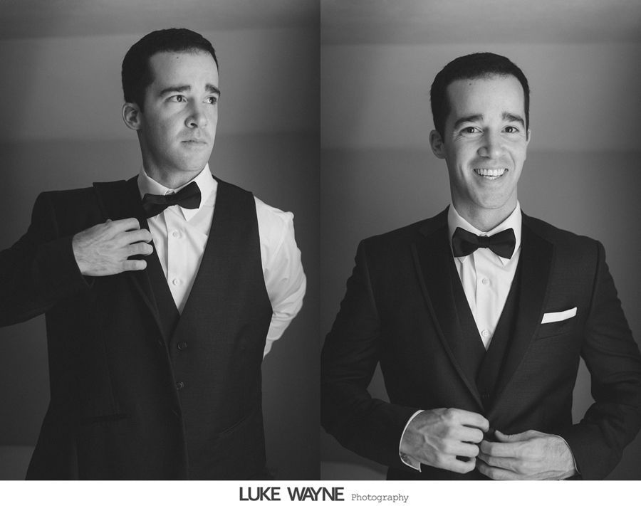 Man in a tuxedo adjusts his bow tie, smiling. Black and white, indoor setting.