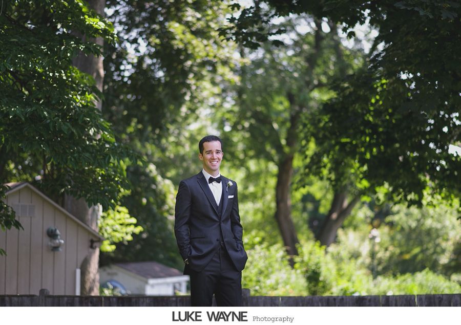 Groom in black tuxedo smiles, standing outdoors near trees and a small building.