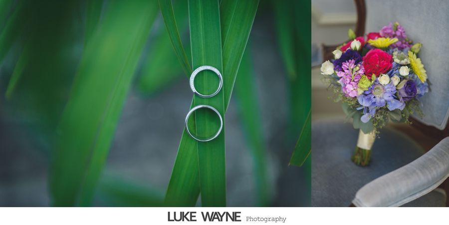 Wedding rings rest on a green leaf; a colorful bouquet is beside them.