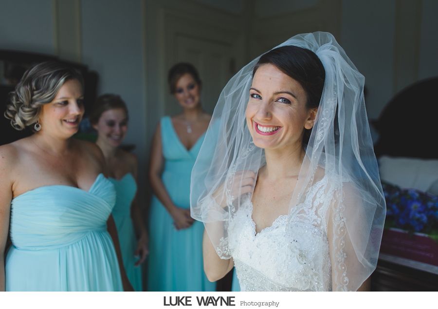 Bride in a white dress and veil smiles, surrounded by bridesmaids in teal dresses in a room.