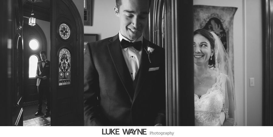 Groom smiles at bride in doorway. She peeks, smiling, wearing a wedding gown and veil. Formal setting.