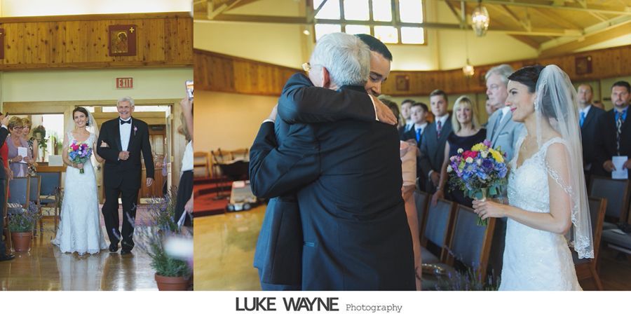 Bride and groom at altar during wedding ceremony. Father of the bride hugs the groom as the bride smiles, holding flowers.