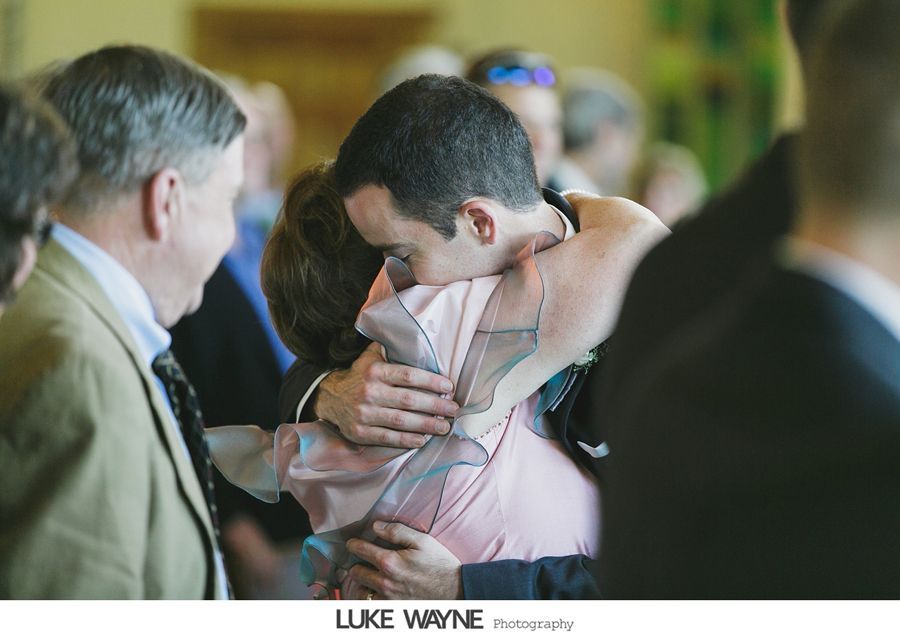 Man hugging a woman, both are embraced and emotional. Other people in the background, a light-filled setting.