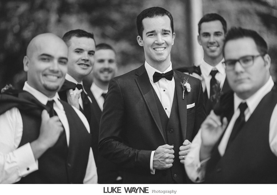 Groom and groomsmen in black tuxedos smiling outdoors. Groom holds his jacket, others wear vests.