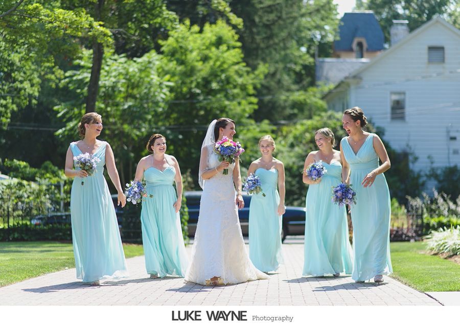 Bride with bridesmaids in turquoise dresses laughing on a brick path, with greenery and a light blue house in the background.