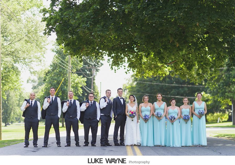 Wedding party stands in road. Groomsmen in navy vests, bridesmaids in turquoise dresses, holding bouquets. Outdoor setting.