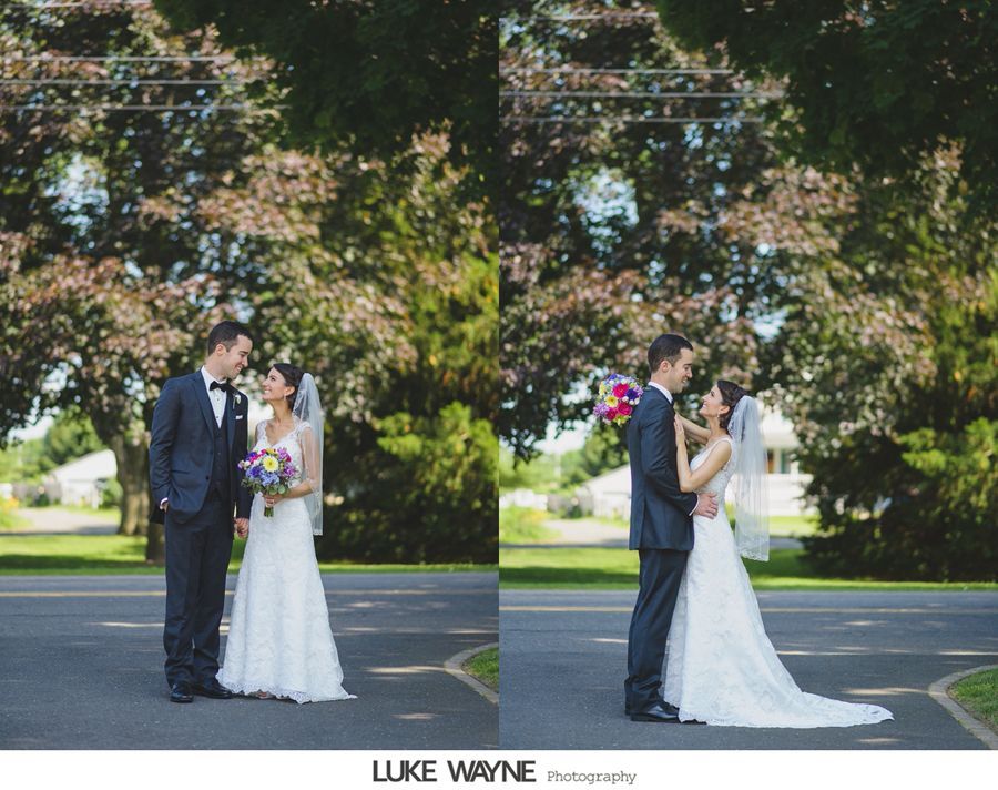 Bride and groom embrace on a road. She wears a white dress, he wears a dark suit. Trees in the background.