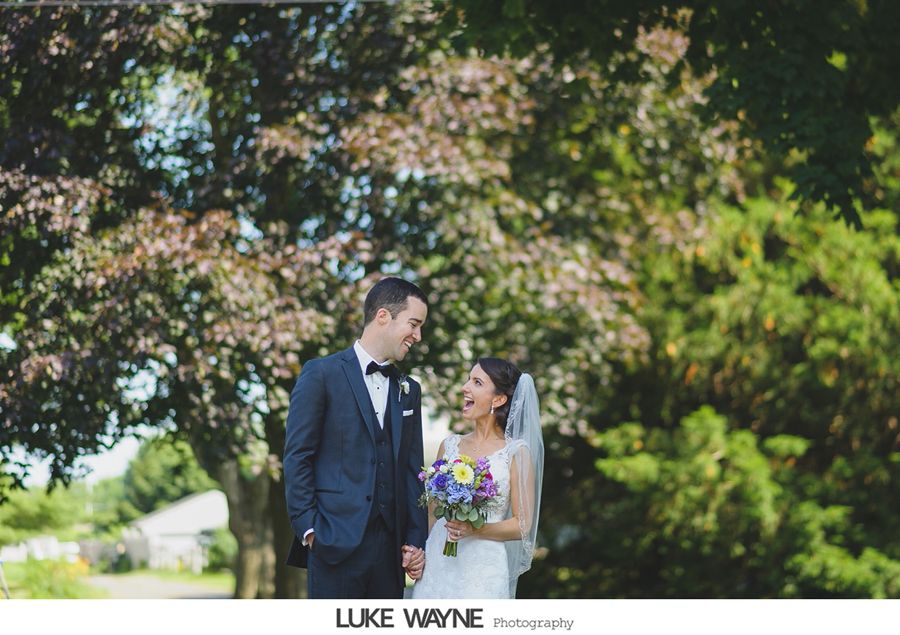 Couple holding hands, smiling at each other, in front of leafy trees on a sunny day. The bride holds flowers.