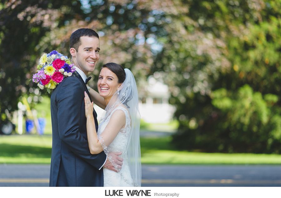 Newlyweds, smiling, embrace outdoors. Man in suit holds bouquet. Woman in dress and veil.