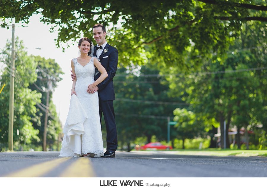 Wedding couple posing in the middle of a tree-lined street; the bride wears white gown; the groom wears a tuxedo.