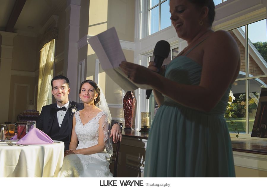 Woman in teal dress gives speech at a wedding. Couple seated at table smiles. Window in background.