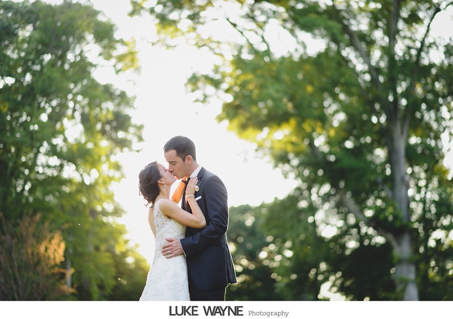 Newlyweds embrace, kissing outdoors with sunlight filtering through trees.