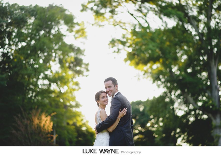 Bride and groom embrace outdoors, surrounded by trees, with sunlight shining between them.