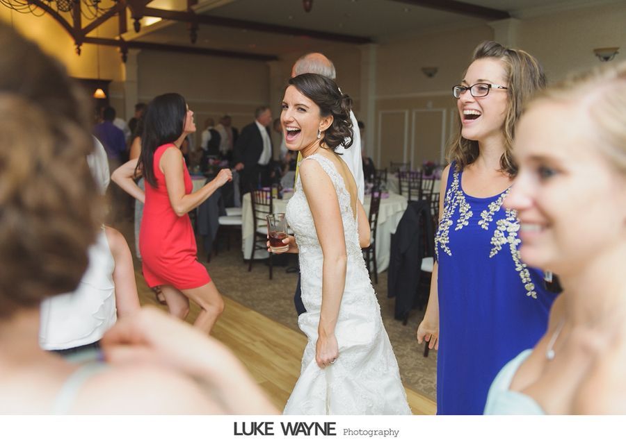 Bride dances joyfully at a wedding reception; guests smile and dance in a room with tables.