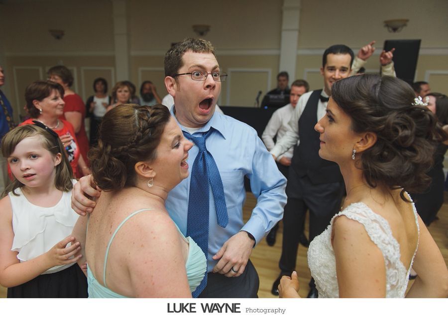 Wedding reception: Man with surprised expression between two women, dancing. Other guests in background.