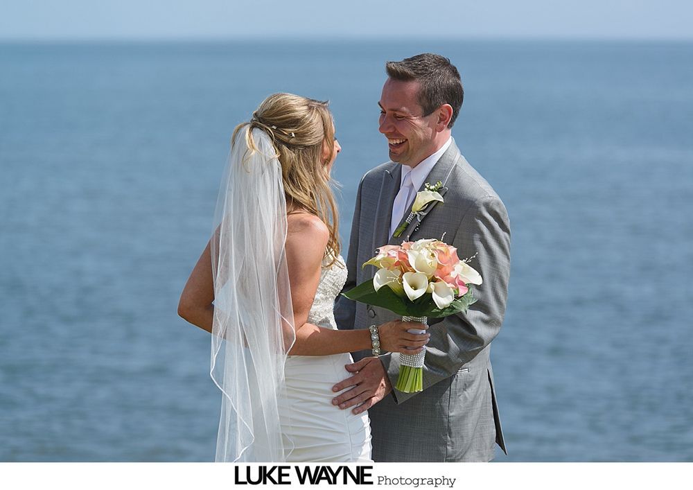 Bride and groom smile at each other, beside ocean. Bride in white gown, groom in grey suit, holding flowers.