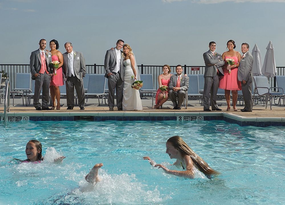 Wedding party by a pool; two girls splash in the water. Wedding party wears formal attire. Sunny outdoor setting.
