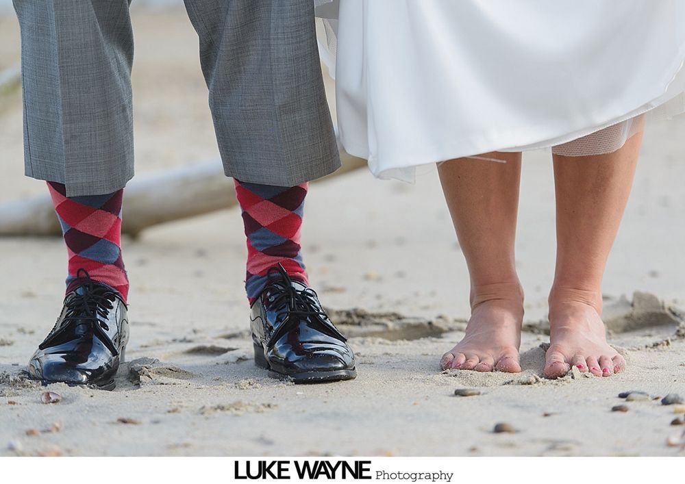 Man in argyle socks and dress shoes stands next to barefoot person in a white dress on a sandy beach.
