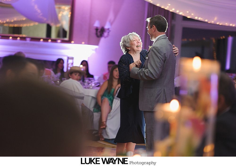 A man and a woman in formal attire dance at a wedding reception; a candlelit table is in the foreground.