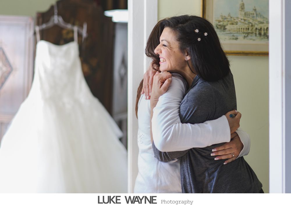 Bride and woman embrace, wedding dress hangs in background. Sunlight, happy expressions.