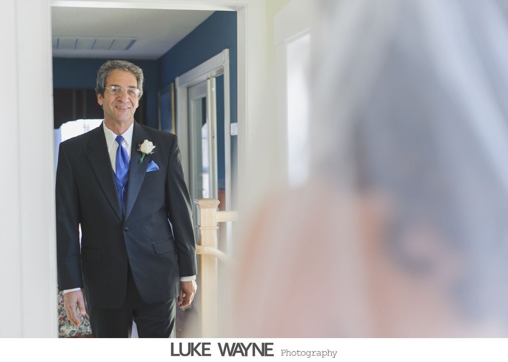 Groom smiles at bride through doorway. He wears a black tuxedo with blue tie and pocket square.