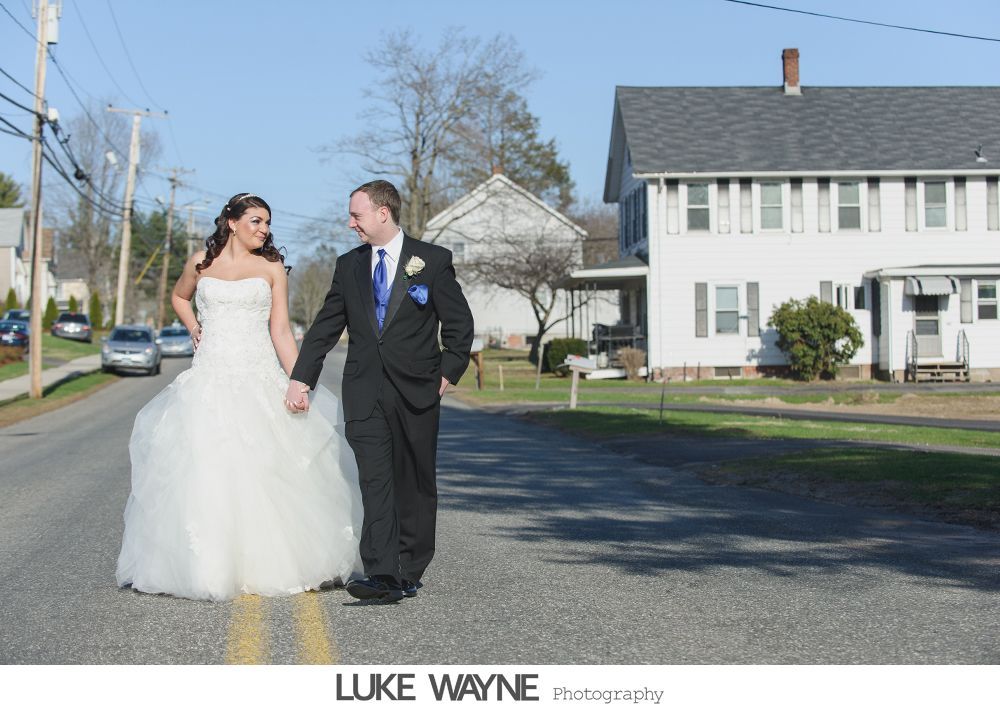 Bride and groom walking in road, holding hands; smiling; near white house.