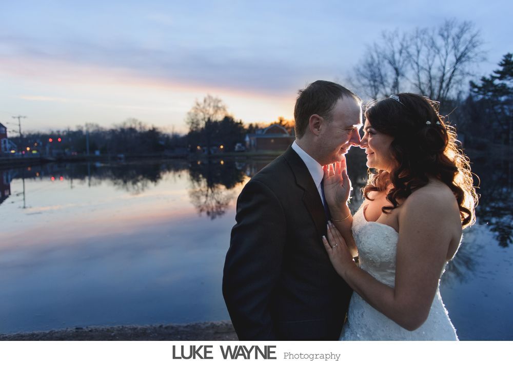 Couple in wedding attire embrace by water at sunset.