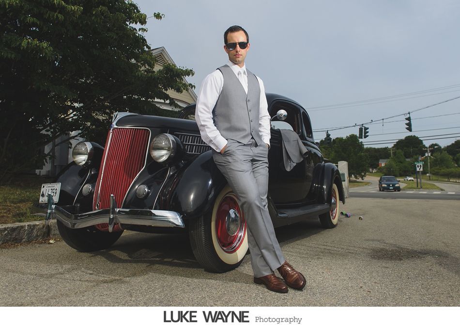 Man in grey suit and sunglasses leans against a classic black car with red grill in an outdoor setting.