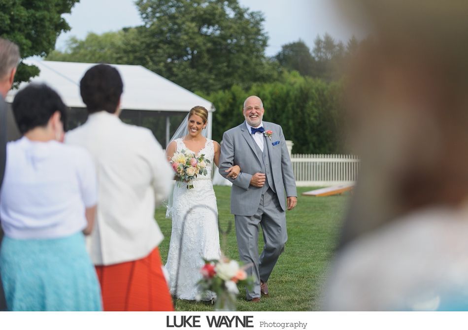 Bride and her father walk down the aisle at an outdoor wedding. Bride smiles, holding a bouquet. Father in a gray suit smiles.