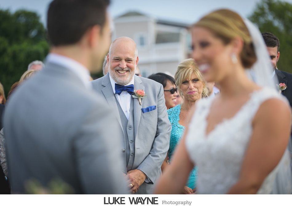 Groom and bride at altar; happy father of the bride smiles. Outdoor ceremony, white dress, grey suit.