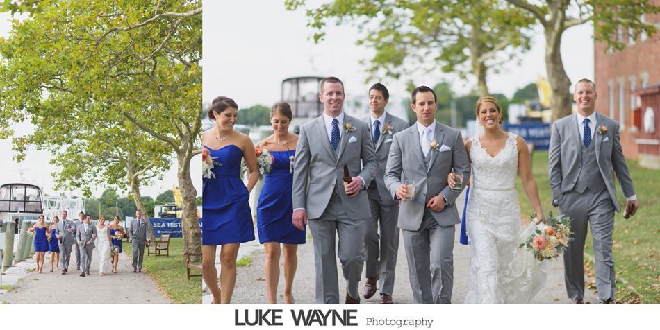 Wedding party walking down a path, harbor setting. Blue dresses and gray suits. Bride in white dress, holding bouquet.