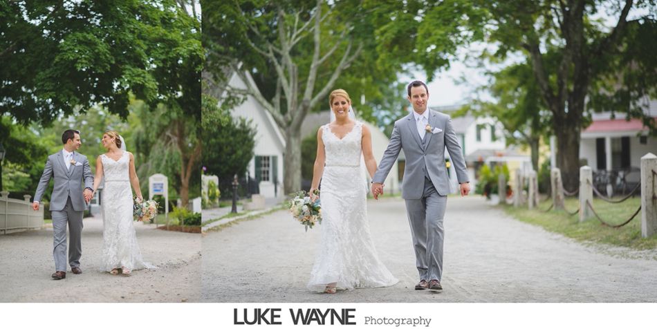 Newlyweds walking hand in hand on a gravel path, smiling, trees and white buildings in background.