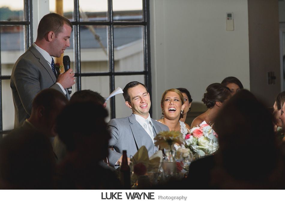 Man giving a speech at a wedding, groom and bride laughing at the table.