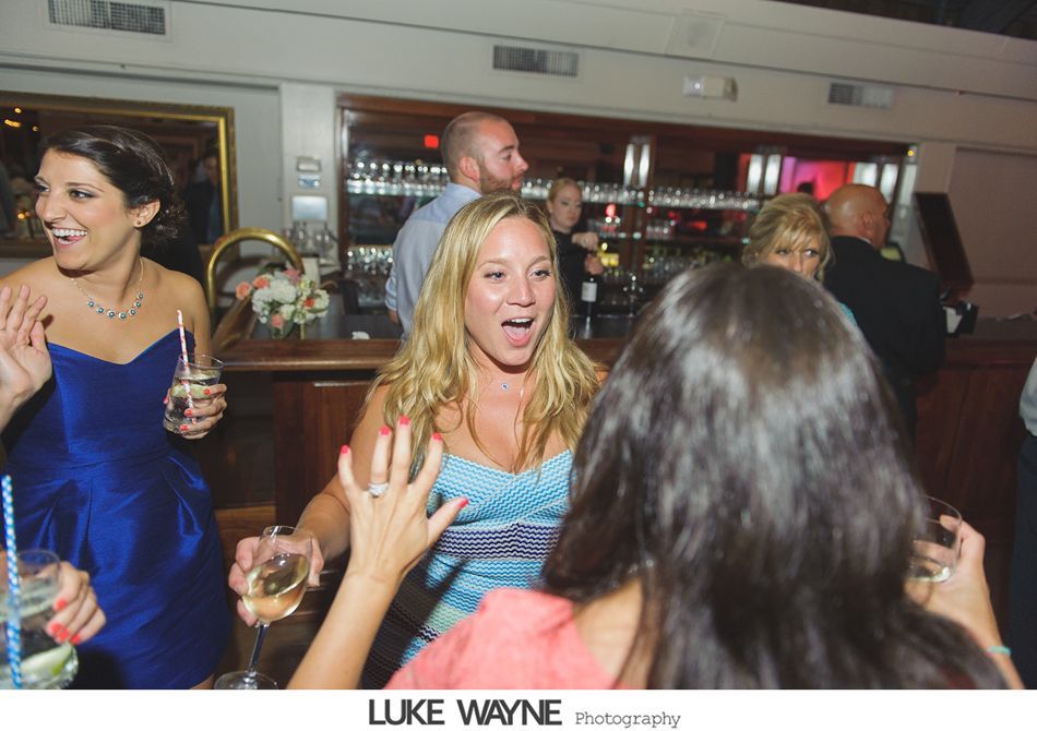 People at a party celebrating, woman with arms raised, holding drink. Others in evening wear near bar.