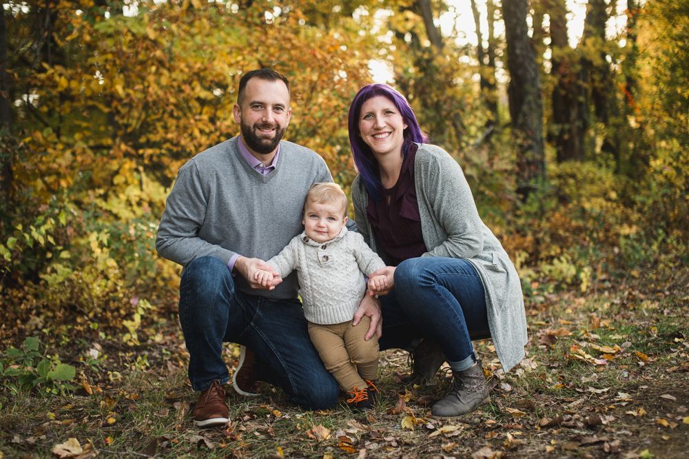 Family of three kneels in a forest, holding hands. Man wears gray sweater, woman has purple hair, child in cable knit. Autumn leaves.