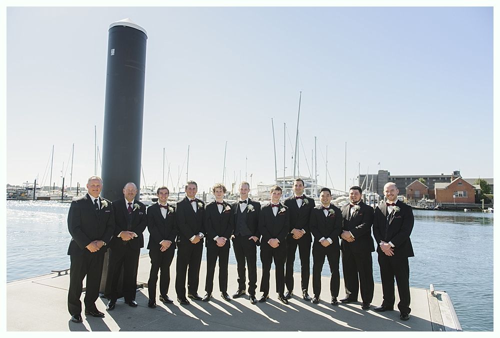 Men in tuxedos stand on a dock in front of sailboats and a tall, black pillar.