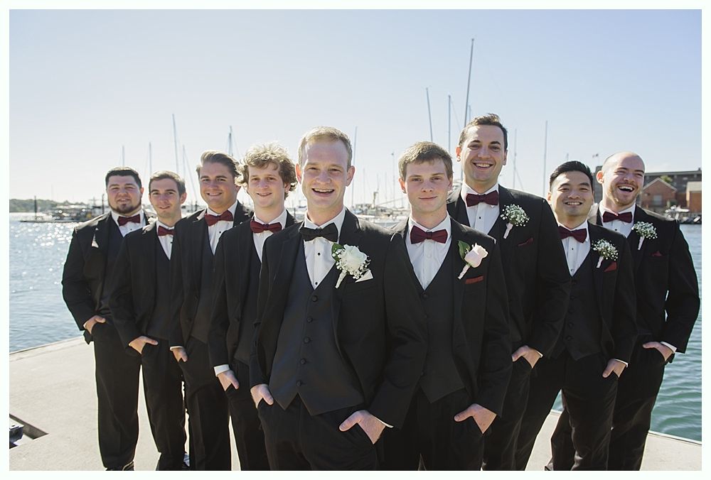 Groomsmen in black tuxedos with burgundy bow ties pose on a dock with a harbor in the background.
