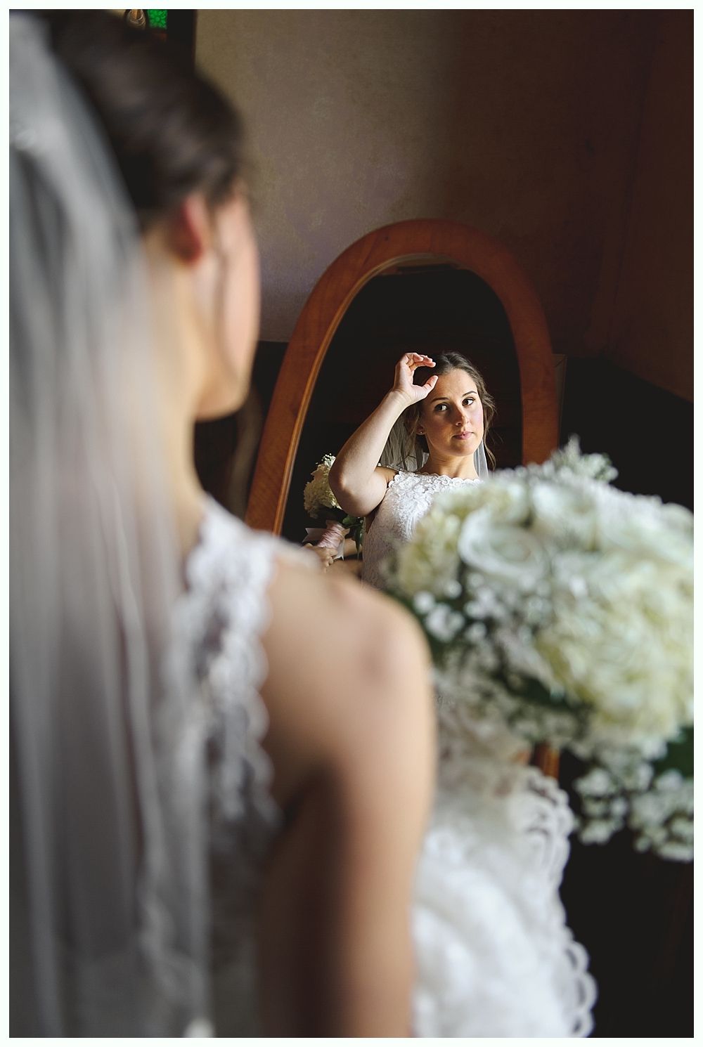 Bride in a white dress with veil, looking at herself in a wooden-framed oval mirror, holding a bouquet.-