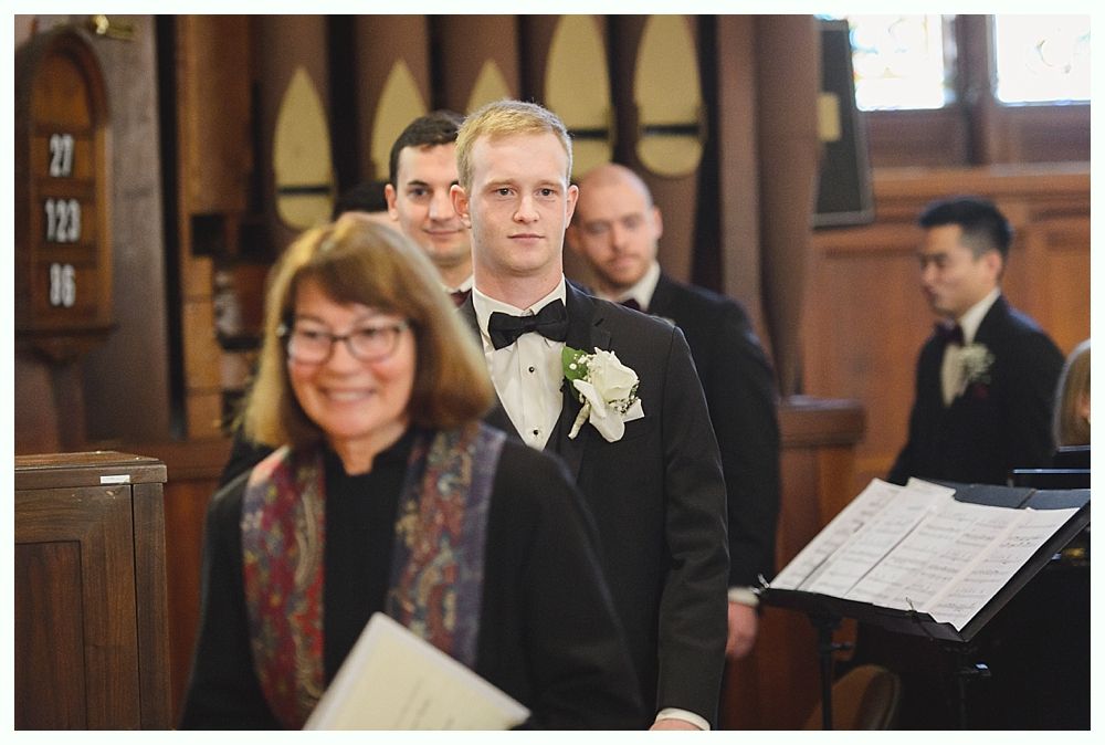 Groom in a tuxedo smiles, walking down a church aisle with his groomsmen, led by a smiling officiant.