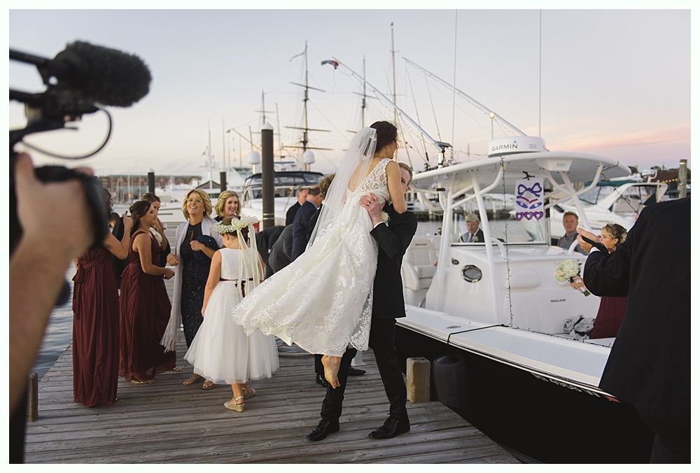 Groom carries bride near a boat at dock, surrounded by wedding party. Evening sky, boat, formal attire.