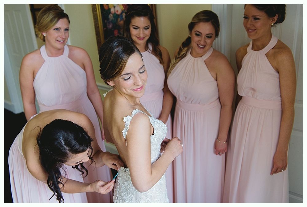 Bride in a white dress being helped by a bridesmaid, surrounded by other bridesmaids in pink dresses.