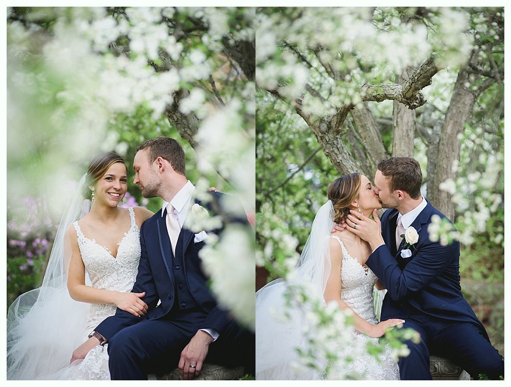 Wedding couple seated under blossoming tree; woman smiles, man kisses; outdoor setting.