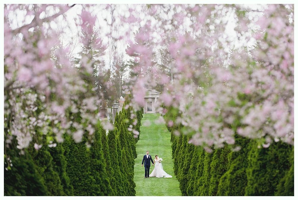 Bride and groom walk down a grassy path lined with evergreen trees and pink flowering trees.