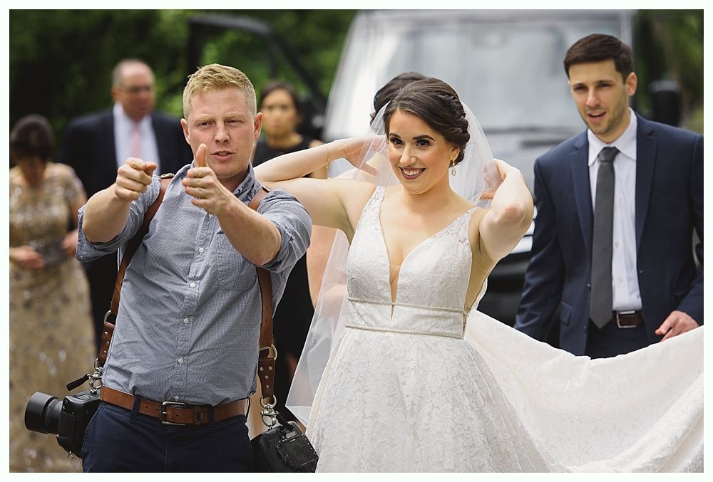Photographer giving thumbs up to bride adjusting veil, with man in suit and wedding party in background.